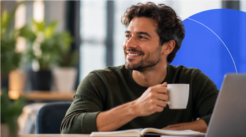 Homem sorridente segura uma xícara de café diante de um notebook e de um caderno aberto.