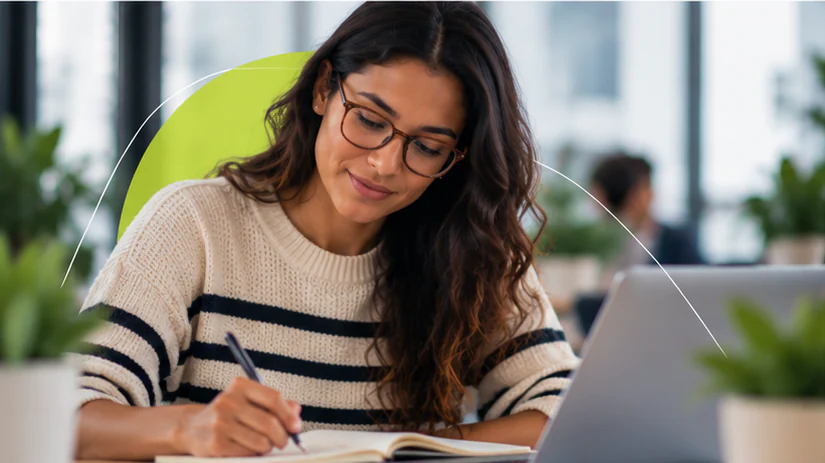 Mulher de óculos e suéter listrado escreve em um caderno ao lado do notebook.