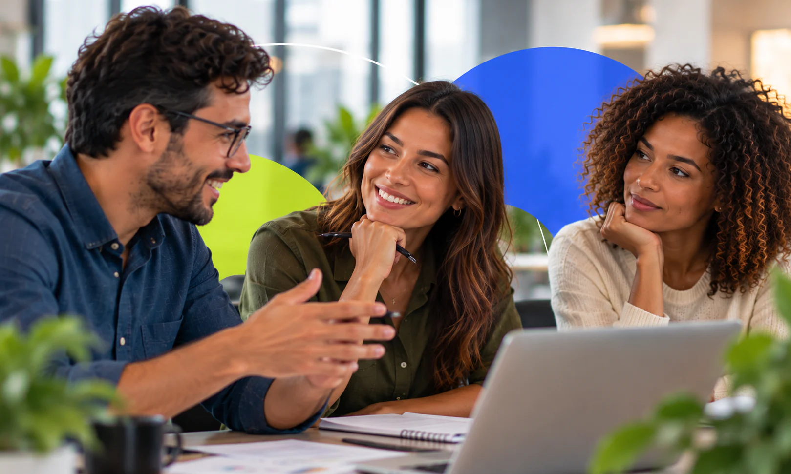 Três colegas conversam e sorriem ao redor de uma mesa com notebook e cadernos.