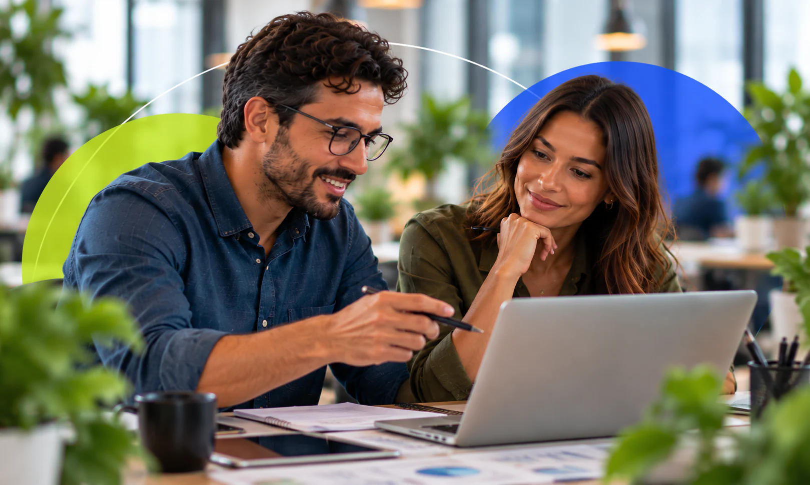 Homem aponta com uma caneta para o notebook enquanto uma colega acompanha sorrindo.