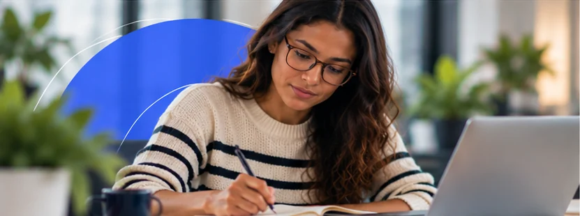 Mulher de óculos faz anotações concentrada, com o notebook à frente e plantas ao redor.