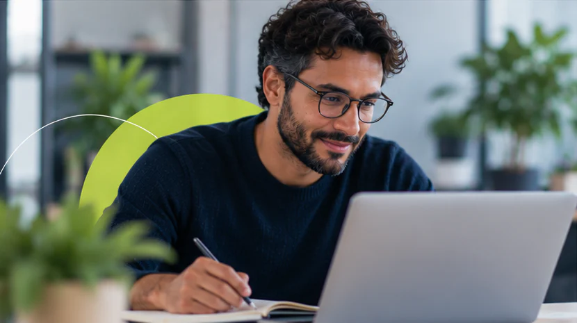 Homem de óculos faz anotações em um caderno ao lado do notebook.