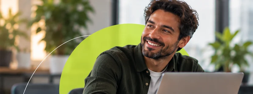 Homem de camisa verde sorri olhando para o lado, sentado diante do notebook.