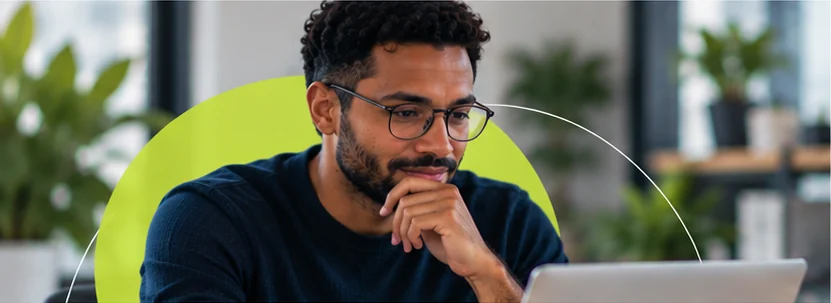 Homem de óculos, com a mão no queixo, observa a tela do notebook com expressão serena.